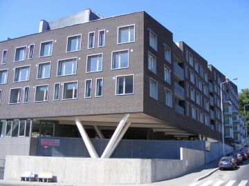 Moderní vícepatrová obytná budova s tmavou fasádou, velkými okny a neobvyklou podperou, vedle zaparkovaných aut na ulici.