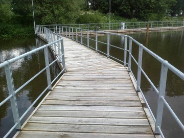 Dřevěná lávka s kovovým zábradlím přes klidnou vodní hladinu, obklopená zelení.