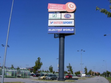 Obchodní centrum OC Géčko se značkami Nákupní centrum, Deichmann, C&A, Intersport, Electro World na parkovišti za slunečného dne.
