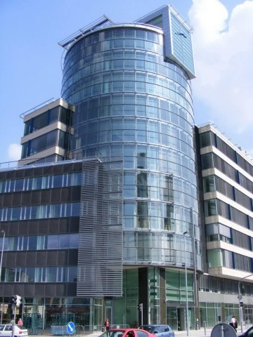 Moderní prosklená kancelářská budova Trianon s válcovou věží pod modrou oblohou.