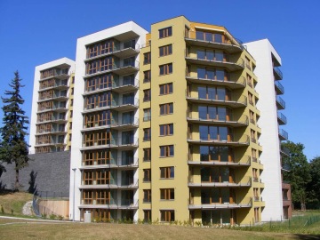 Moderní bytový dům Vokovice se skleněnými balkony a žlutou fasádou pod modrou oblohou.