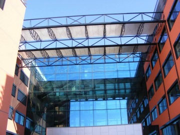 Moderní budova společnosti Skanska s prosklenými stěnami a oranžovými zdmi pod průhledným přístřeškem ze skla a kovu.