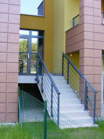 Moderní budova Vokovice s venkovním schodištěm a hnědou fasádou, obklopená zeleným trávníkem a plotem.