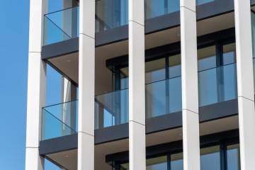 Moderní budova s balkony, skleněnými zábradlími a bílými vertikálními panely proti modré obloze.