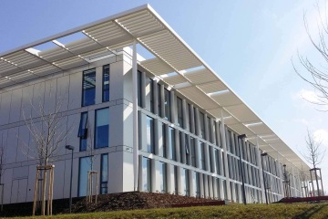 Moderní budova Laserové centrum ELI s prosklenou fasádou a bílými sloupky pod modrou oblohou.