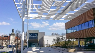 Moderní budova Laserové centrum ELI s prosklenými okny a bílou střechou, v pozadí historická věž kostela, modrá obloha s oblaky.