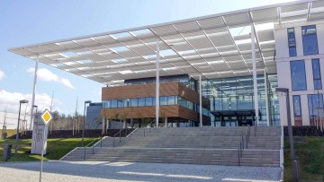 Moderní budova Laserové centrum ELI se skleněnou fasádou a dřevěnými prvky, schody vedoucí k hlavnímu vstupu, pergola nad budovou.