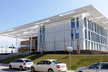 Moderní budova Laserové centrum ELI s bílou fasádou a velkým stínovým přístřeškem, v popředí zaparkovaná auta na ulici.