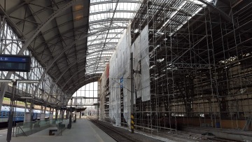 Železniční nádraží s opravenými nástupišti a kovovou konstrukcí pokrytou ochrannou plachtou.