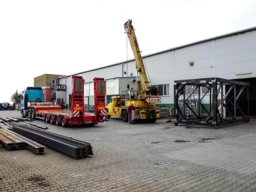 Žlutý jeřáb manipulující s ocelovou konstrukcí pod sila vedle nákladního vozu na firemním dvoře.