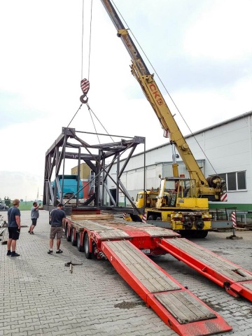 Vyložení velké kovové konstrukce pod sila z nákladního auta pomocí žlutého jeřábu, pracovníci sledují proces.