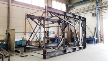 Kovová konstrukce pod sila ve výrobní hale, obklopena pracovními stoly a nástroji.