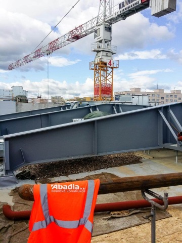 Červený jeřáb na staveništi, oranžová vesta Abadia na zábradlí, ocelové konstrukce na pozadí.