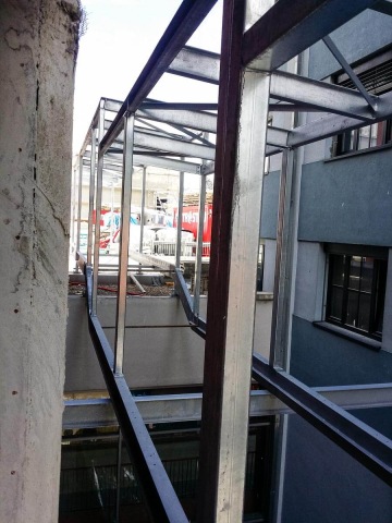 Konstrukce kovového mostu mezi dvěma budovami s pohledem na okolní městskou architekturu.