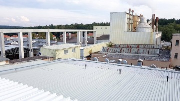 Průmyslový komplex s budovami OK haly a komíny, obklopený stromy a nízkými stavbami.