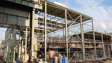 Stavba ocelové konstrukce průmyslové budovy OK haly s potrubím a pracovními dělníky, modrobílá obloha v pozadí.