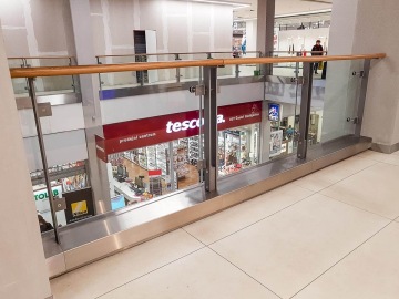 Obchodní centrum s prosklenými zábradlími, výhledem na prodejnu Tesco se zákazníky a zbožím.