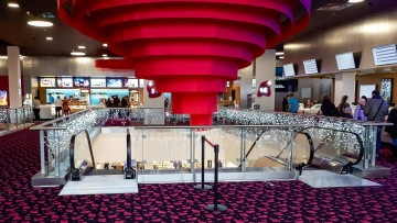 Moderní interiér kina s červeným spirálovitým stropem, schody, zábradlí zdobené světly a zákazníci u pokladen.