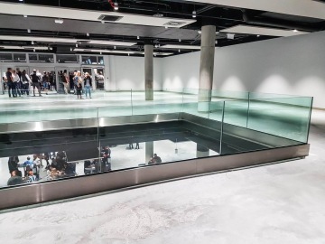 Moderní výstavní prostor O2 Universum Praha s prosklenými zábradlími a betonovou podlahou, lidé stojící v pozadí.