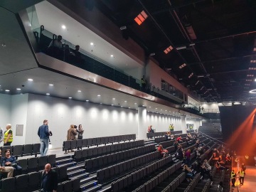 Poloprázdný moderní sál O2 Universum Praha s lidmi sedícími na tribunách a několik stojících osob; k osvětlení prostoru přispívají zářivky.