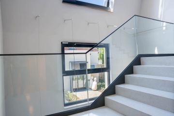 Skleněné zábradlí moderního schodiště Chateau Troja Residence, okno s výhledem na zahradu a sochy venku.