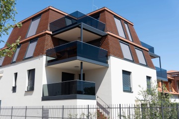 Moderní víceúrovňová budova Chateau Troja Residence s tmavými balkóny a dřevěnými lamelami na fasádě, jasně modrá obloha.