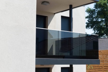 Moderní balkon se skleněným zábradlím u bílé budovy Chateau Troja Residence, na pozadí stromy a střecha.