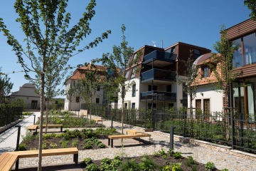 Moderní bytové domy Chateau Troja Residence s dřevěnými a skleněnými prvky, obklopené zahradou a lavičkami pod modrou oblohou.