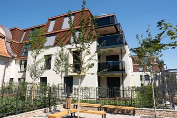 Moderní bytový dům Chateau Troja Residence s prosklenými balkony, kovovým plotem a lavičkami na dlážděném dvoře, obklopený mladými stromy.