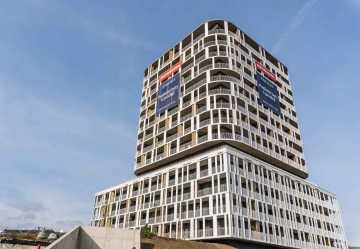 Moderní obytná budova ve Čtvrti Zahálka s komplexním designem fasády a reklamními cedulemi na prodej bytů pod modrou oblohou.