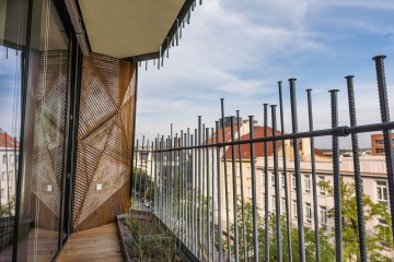 Balkón s kovovým zábradlím v Dům Komunardů XXXV a dekorativní dřevěnou stěnou, výhled na město s červenými střechami pod modrou oblohou.