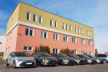 Řada zaparkovaných automobilů před dvoupatrovou budovou v jasných barvách pod modrou oblohou.