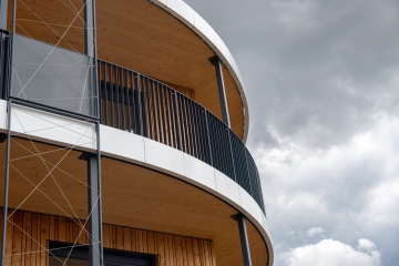 Moderní kruhový balkón s dřevěným obložením a kovovými prvky pod zataženou oblohou.