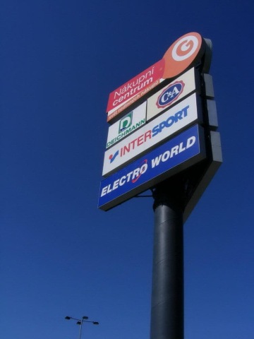 Reklamní poutač OC Géčko nákupního centra se značkami Deichmann, C&A, Intersport a Elektro World na modré obloze.