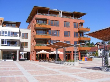 Moderní bytové domy Dolní Břežany s terasami na slunném náměstí, stoly se slunečníky v popředí.