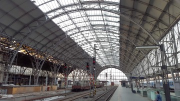 Železniční stanice s klenutou skleněnou střechou a prázdnými kolejemi, na perónu sedí žena.