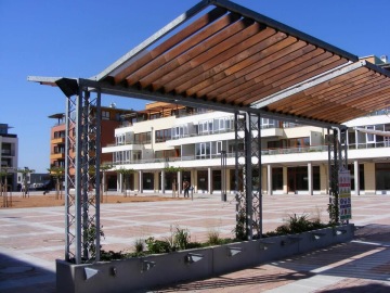 Moderní náměstí Dolní Břežany s pergolou a obytnými domy v pozadí pod jasnou modrou oblohou.