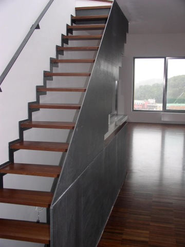 Dřevěné vnitřní schodiště s kovovým zábradlím v moderním interiéru s velkými okny.