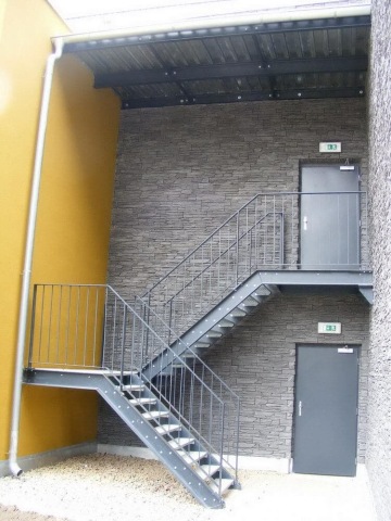 Ocelové schodiště vedle kamenné zdi, s dveřmi na horní i dolní úrovni, nad podlahou z jemného štěrku.