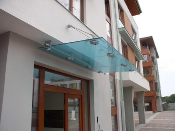 Skleněná stříška nad venkovním vchodem moderní budovy Dolní Břežany se dřevěnými dveřmi.