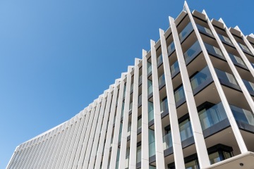 Moderní budova s elegantními bílými lamelami a skleněnými balkony proti modré obloze.