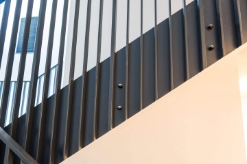 Moderní zábradlí s vertikálními ocelovými pruty v interiéru Rezidence MAISON Ořechovka, vedle okna s černými žaluziemi.