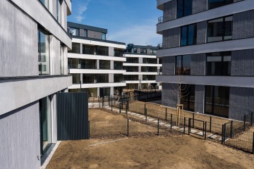 Moderní bytové domy Rezidence MAISON Ořechovka s prosklenými balkóny, ploty a minimální stromy na dvoře za slunečného počasí.