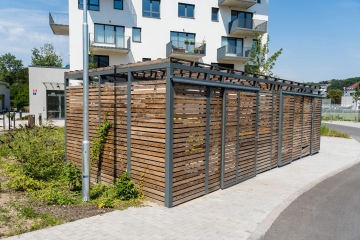 Dřevěný přístřešek s kovovou konstrukcí u moderní budovy, vedle vydlážděného chodníku, obklopený zelení.