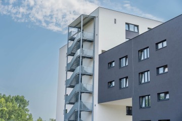 Moderní bytový dům AFI Home Kolbenova s venkovním kovovým schodištěm na pozadí modré oblohy.