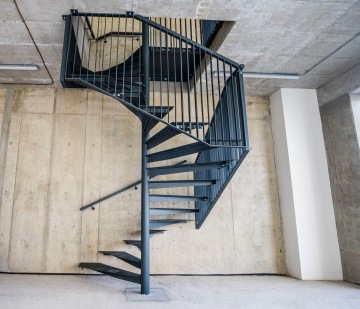 Moderní černé kovové točité schodiště vedoucí k hornímu patru AFI Home Třebešín v betonovém interiéru.