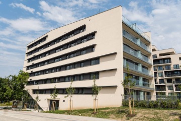 Moderní bytová budova AFI Home Třebešín s několika patry, velkými prosklenými balkony a světlou omítkou, na pozadí je obloha s mraky.