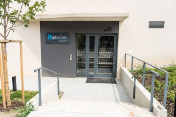 Vchod do budovy AFI Home Třebešín s dveřmi a cedulí na zdi, schody a zábradlí.