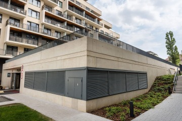 Moderní bytový dům AFI Home Třebešín s betonovou terasou, parkovacím vchodem a zelení podél cesty.