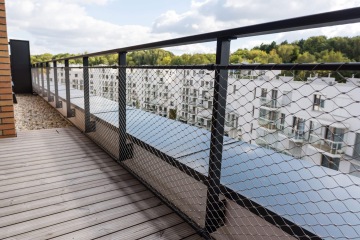 Balkón YIT Parvi Cibulka s dřevěnou podlahou a kovovým zábradlím s ochrannou sítí, výhled na bílé moderní budovy a zeleň.